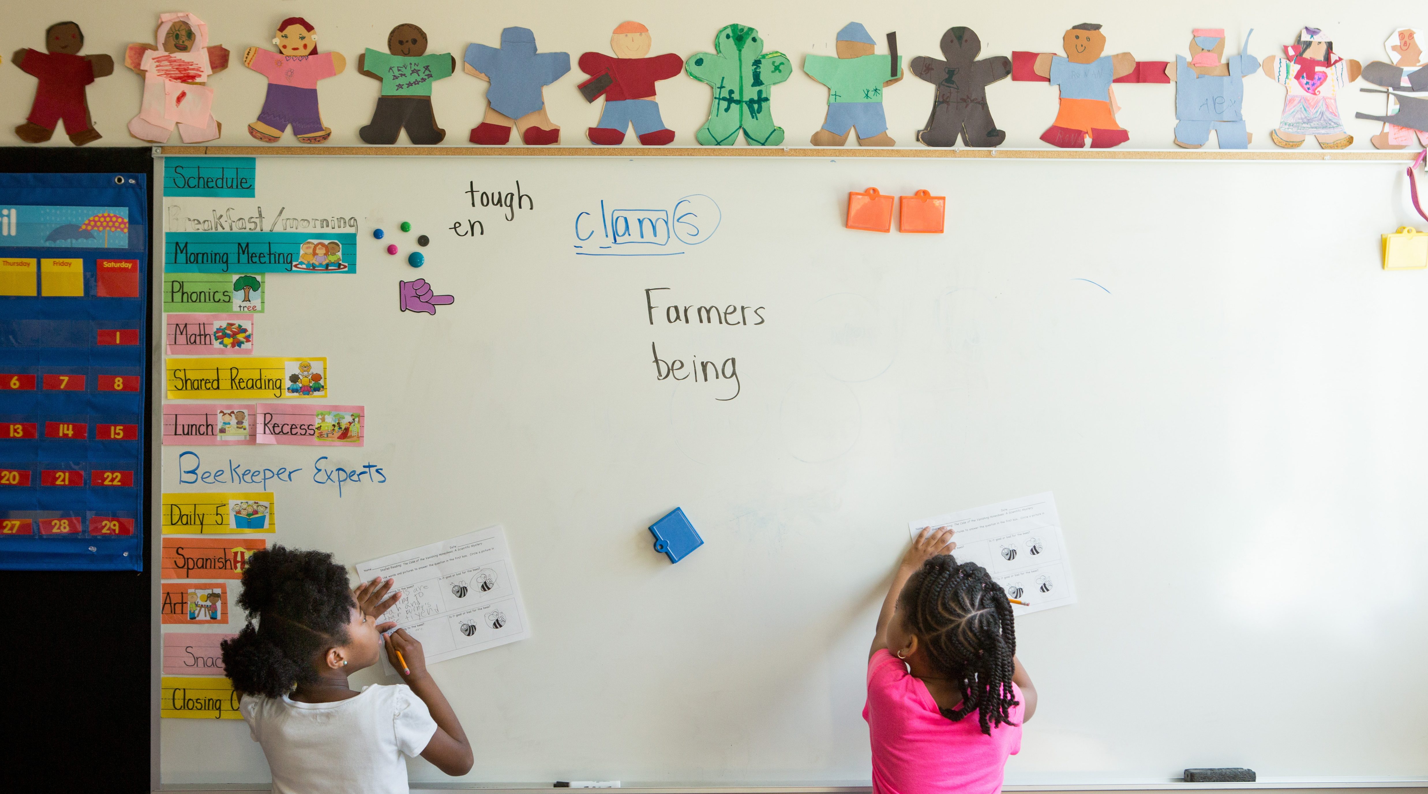 Two students complete worksheets facing a classroom whiteboard.