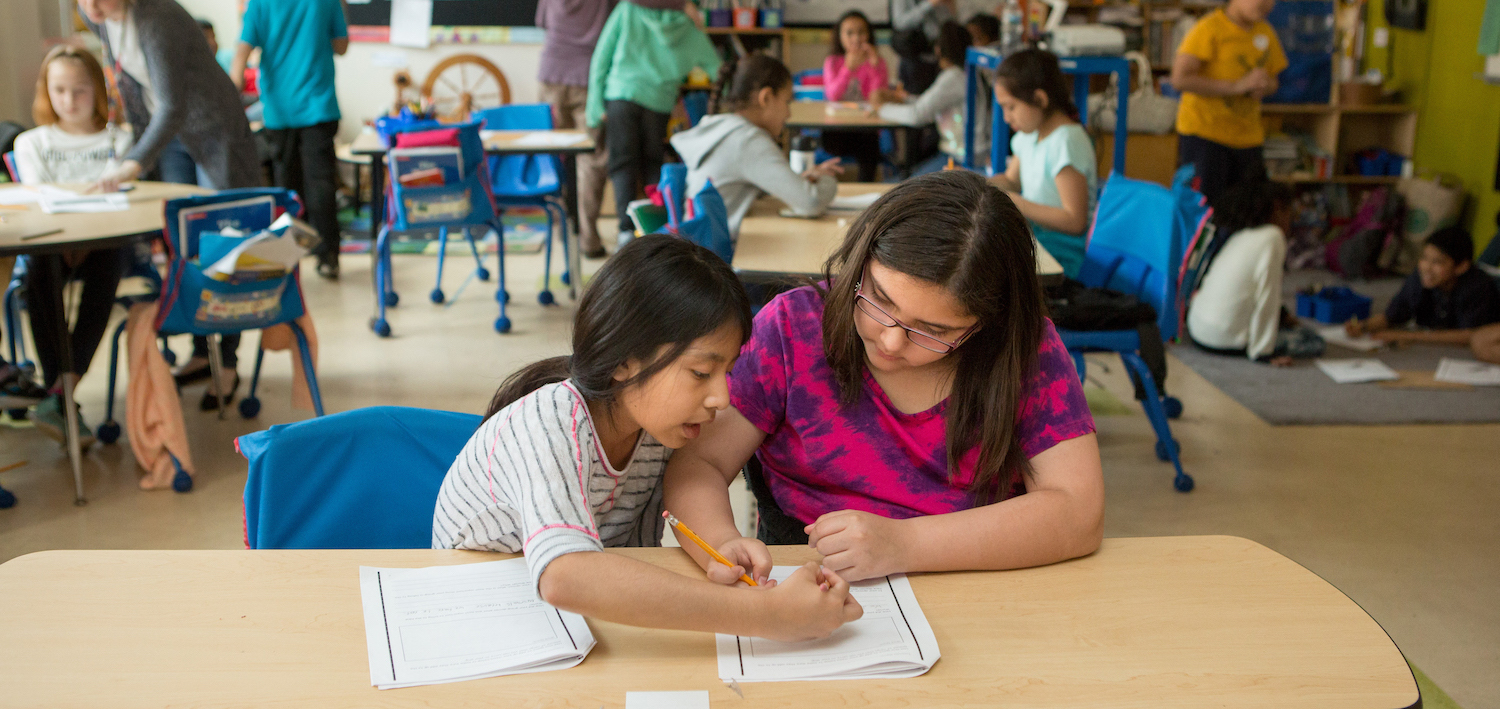 Two students sit side by side in a classroom, completing a worksheet together.